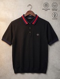 A black short-sleeve polo shirt with a red and navy blue striped collar, hanging on a wooden hanger against a textured gray wall. The shirt features a small embroidered bee logo on the left chest and includes three text callouts in the upper right: 'BREATHABLE KNIT', 'PREMIUM COTTON', and 'SLIM FIT'.