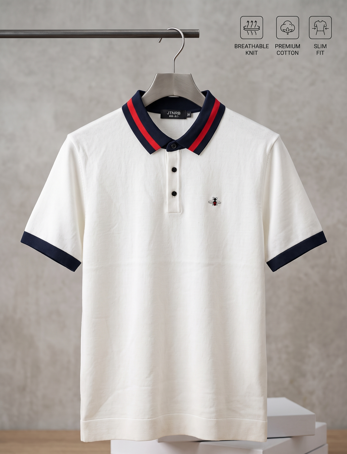 A white short-sleeve polo shirt with navy and red striped collar trim and navy sleeve cuffs, hanging on a hanger against a neutral textured wall. The shirt has a small embroidered logo on the left chest and is displayed on top of stacked white boxes, with three feature icons at the top indicating breathable knit, premium cotton fit, and slim fit.